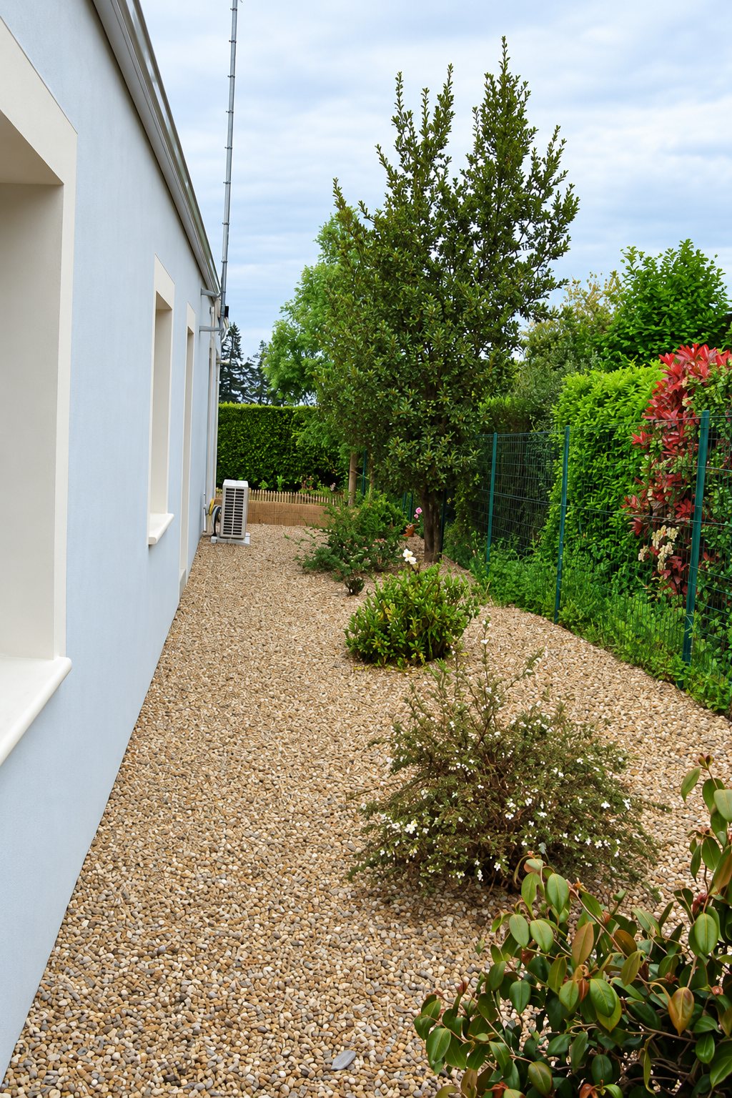 Une allée couverte de gravier longe une maison aux murs bleu clair, bordée d'arbustes fleuris, d'une haie et d'arbres.