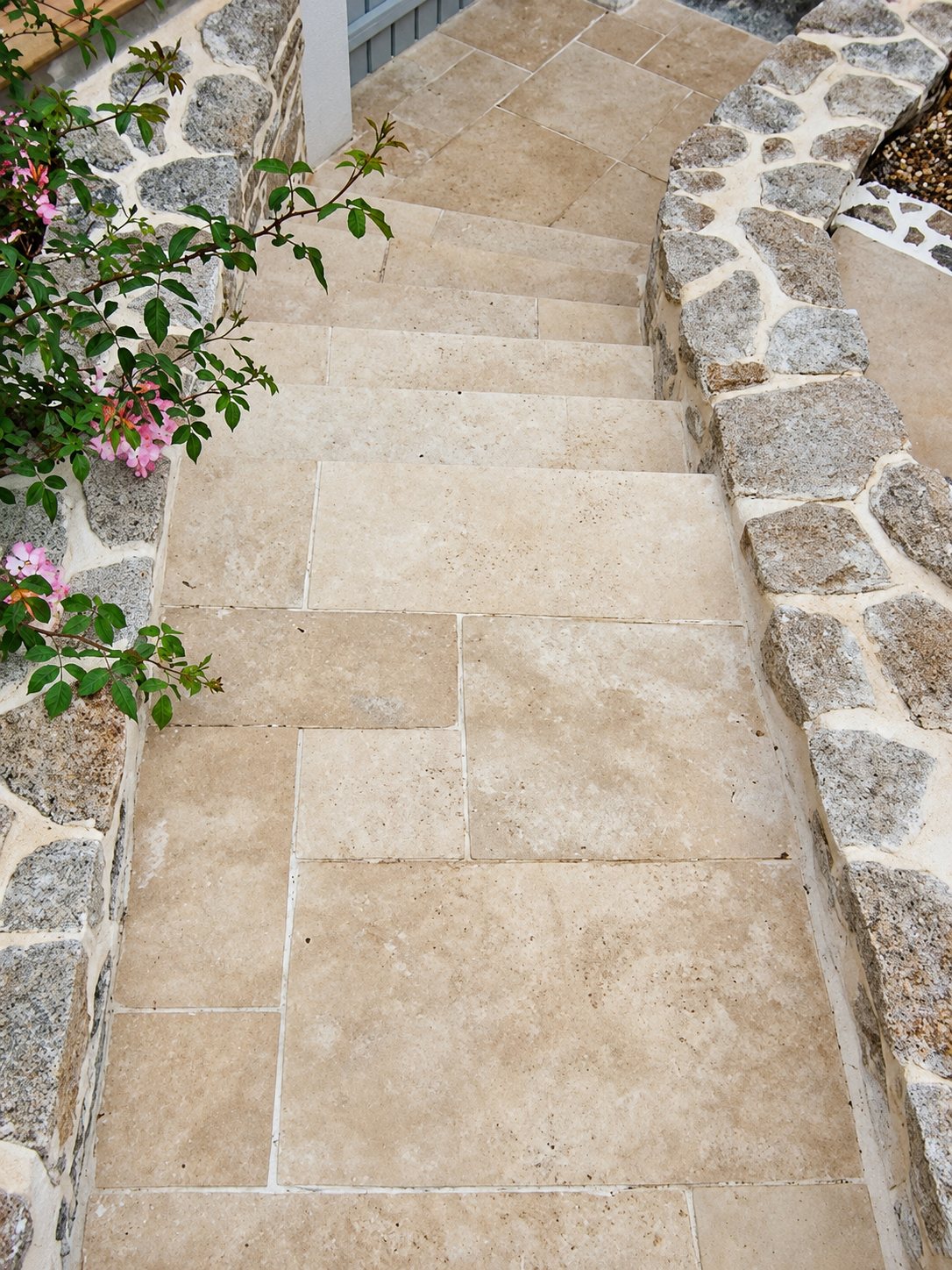 Un escalier extérieur recouvert de grandes dalles de pierre beige, bordé de murets en pierres de granit et de rosiers en fleurs.