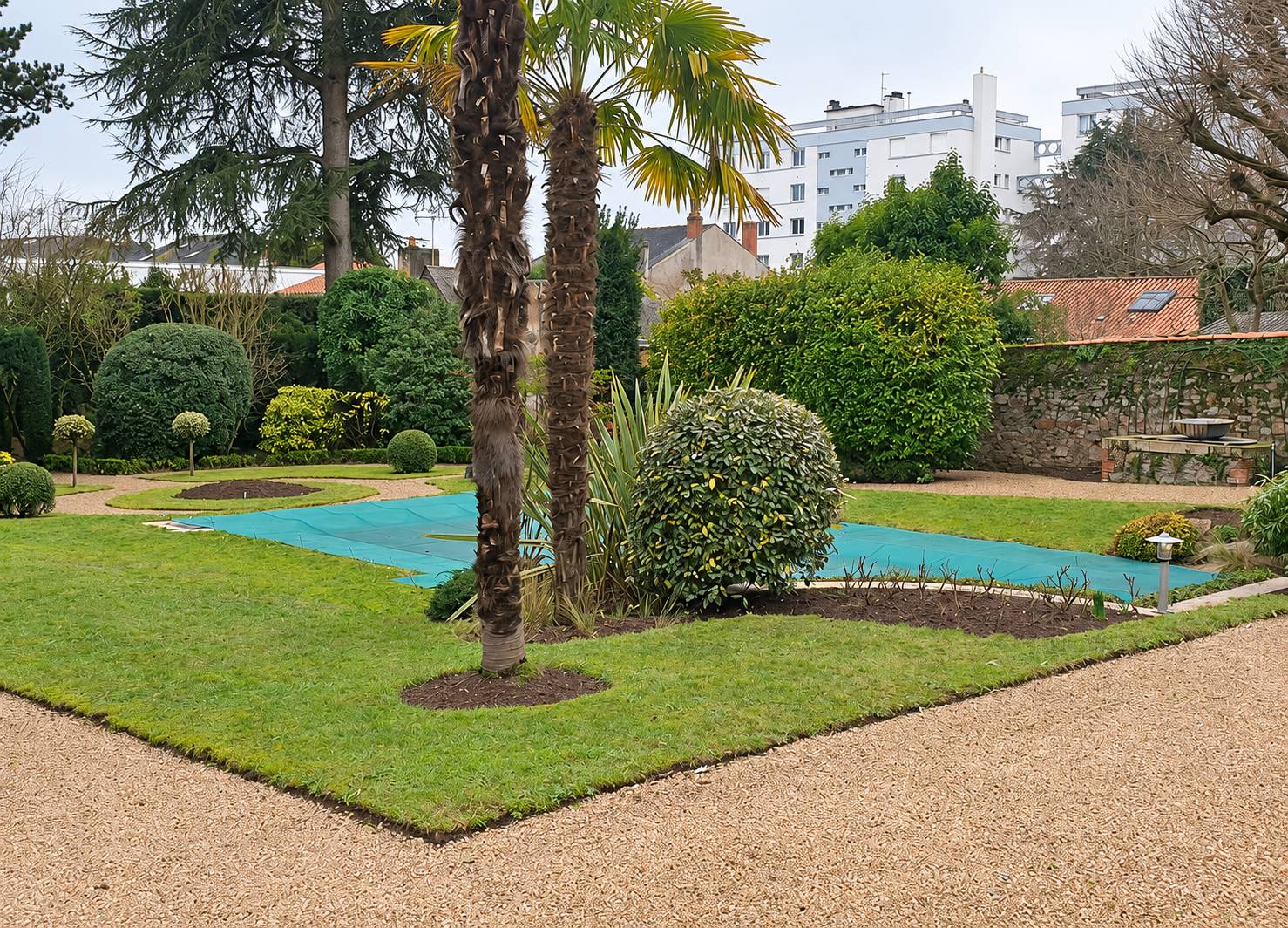 Jardin urbain soigné avec pelouse, palmiers, arbustes taillés en boule et une piscine recouverte d'une bâche verte, bordé d'une allée de gravier.